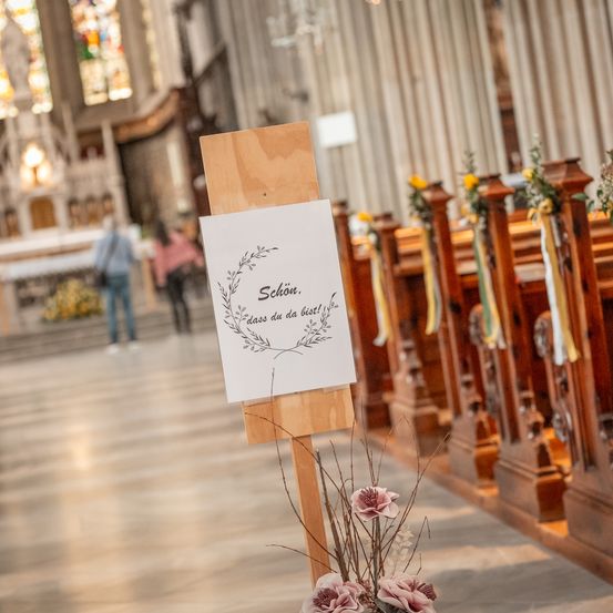 Ein hölzernes Schild mit einer dekorativen Girlande befindet sich in einem Kirchengang, mit zwei Personen, die im Hintergrund gehen. Das Schild trägt die Aufschrift 'Schön, dass du da bist!' Das Kircheninnere zeigt Buntglasfenster und Holzbänke.