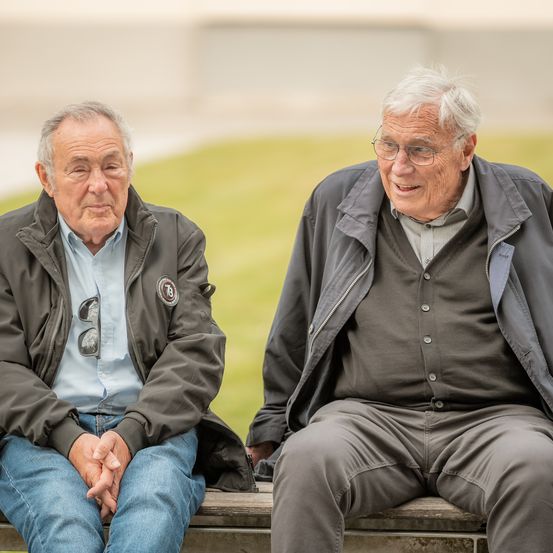 Zwei ältere Männer sitzen auf einer Bank im Freien. Einer trägt eine Jacke mit Logo, eine Brille und ein geknöpftes Hemd. Der andere trägt eine Jacke, eine Brille und ein graues Hemd und lächelt.