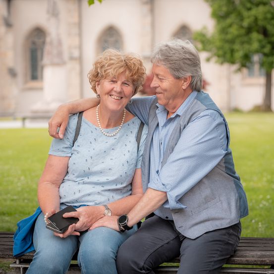Ein älteres Paar, eine Frau mit einer Geldbörse und ein Mann mit seinem Arm um sie herum, sitzt auf einer Parkbank. Hinter ihnen befindet sich ein Gebäude mit Rundbogenfenstern.