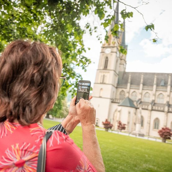 Eine ältere Frau macht ein Foto einer Kirche mit ihrem Handy. Sie steht auf einem Rasen mit einem Baum hinter ihr. Die Kirche hat einen Turm mit einer Uhr.