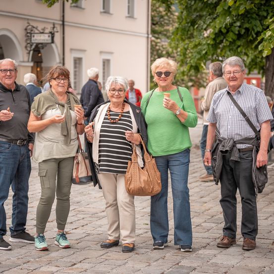 Fünf Menschen stehen zusammen in einem Außenbereich, wahrscheinlich einer Straße. Sie lächeln und scheinen für ein Foto zu posieren. Sie halten Eisbecher in der Hand. Hinter ihnen steht ein Gebäude mit einem Schild.