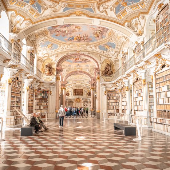 Eine große Bibliothek mit kunstvollen Deckenfresken und hohen Bücherregalen. Besucher sitzen auf Bänken und andere gehen durch die Gänge.