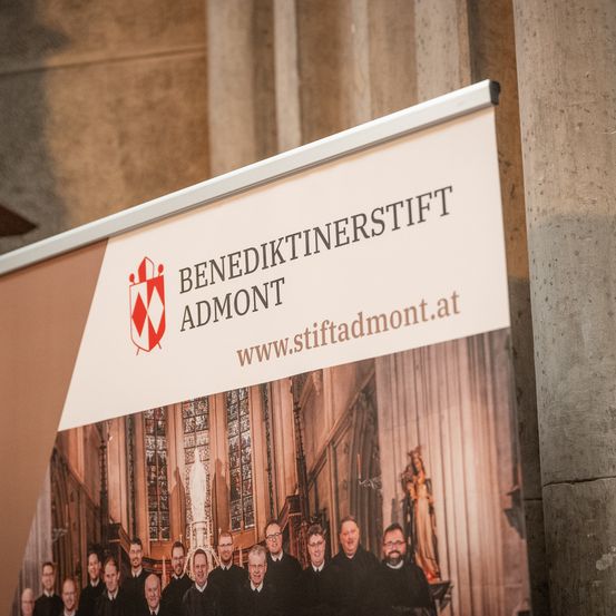 Ein Schild für das Benediktinerstift Admont mit einem Männerchor, einem Logo und einer Website-Adresse, vor dem Hintergrund einer Kathedrale.