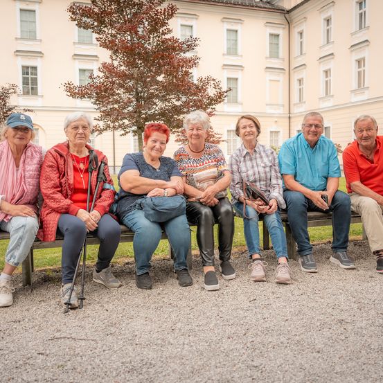 Eine Gruppe älterer Menschen sitzt auf einer Bank vor einem historischen Gebäude, einige tragen Brillen, Hüte und Halsketten. Sie scheinen für ein Foto zu posieren.