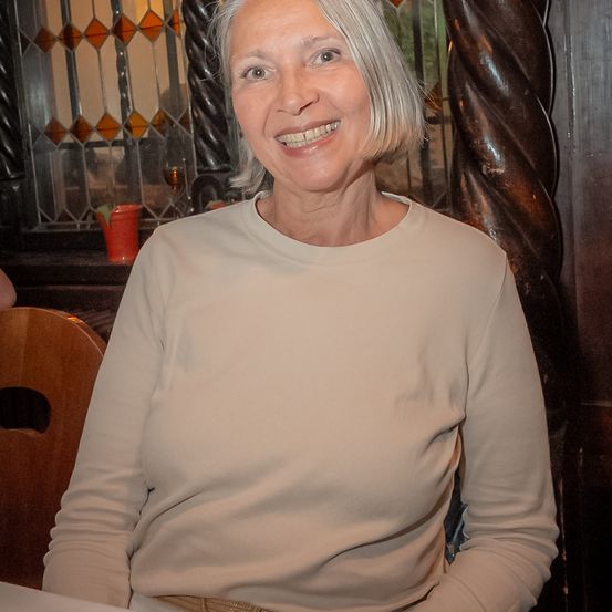 Eine Frau mit kurzen weißen Haaren und einem warmen Lächeln posiert drinnen, trägt ein hellbeiges Langarmshirt. Hinter ihr sind ein Holzstuhl und eine dekorative Holzsäule zu sehen. Im Hintergrund befindet sich ein Buntglasfenster und ein roter Becher auf der Fensterbank.