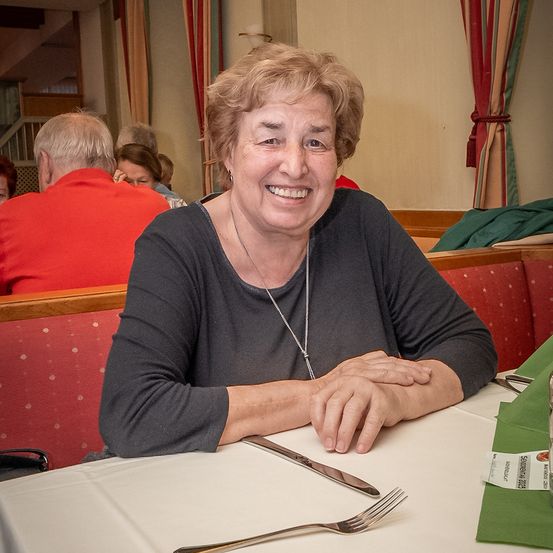 Eine Frau mit kurzen Haaren, die ein graues Shirt und eine Halskette trägt, sitzt lächelnd an einem Tisch in einem Restaurant. Hinter ihr ist ein Mann in einem roten Hemd zu sehen.
