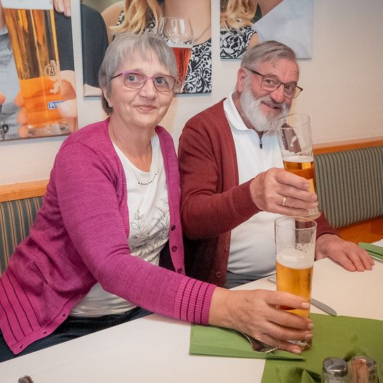 Zwei ältere Menschen, ein Mann und eine Frau, sitzen an einem Tisch, lächeln und stoßen mit Biergläsern an. Die Frau trägt eine Brille und eine rosa Strickjacke, während der Mann einen roten Pullover trägt und einen Bart hat. Hinter ihnen hängen mehrere Bilder an der Wand.