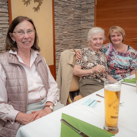Drei Frauen sitzen an einem Tisch. Die Frau auf der linken Seite trägt eine Brille und eine Uhr. Die Frau in der Mitte hat ihren Arm um die andere Frau gelegt. Es gibt ein Glas Bier und eine Karte auf dem Tisch.
