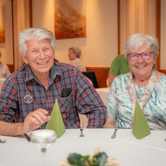Zwei lächelnde ältere Menschen sitzen in einem Restaurant an einem Tisch, sie tragen Brillen und farbenfrohe Hemden. Der Tisch hat grüne Servietten, eine Kerze und Besteck. Im Hintergrund sitzen weitere Personen.