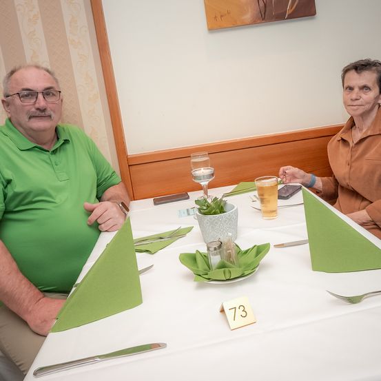 Zwei ältere Erwachsene sitzen lächelnd an einem Tisch mit Getränken und einer Topfpflanze, eine Karte mit der Nummer 73 auf dem Tisch und dekorative Servietten und Besteck.
