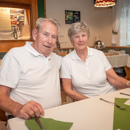Ein älteres Paar sitzt an einem Tisch in einem Restaurant. Sie tragen beide weiße Poloshirts und scheinen für ein Foto zu posieren. Eine grüne Serviette und Besteck sind auf dem Tisch. Hinter ihnen steht ein Tisch mit weißer Tischdecke, einer Flasche und anderen Gegenständen.