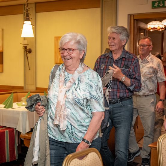 Drei ältere Personen stehen in einem Essbereich. Eine Frau hält ihre Jacke und lächelt. Ein Mann in einem karierten Hemd gestikuliert. Ein anderer Mann in einem Blumenhemd beobachtet. Ein Tisch mit weißer Tischdecke ist in der Nähe.