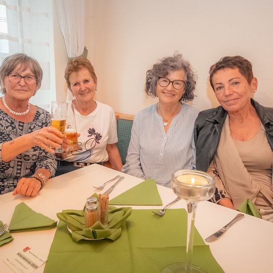 Vier Frauen sitzen an einem Tisch und lächeln für ein Foto. Sie halten Getränke in der Hand, und der Tisch ist mit grünen Servietten, Besteck und Kerzen gedeckt.