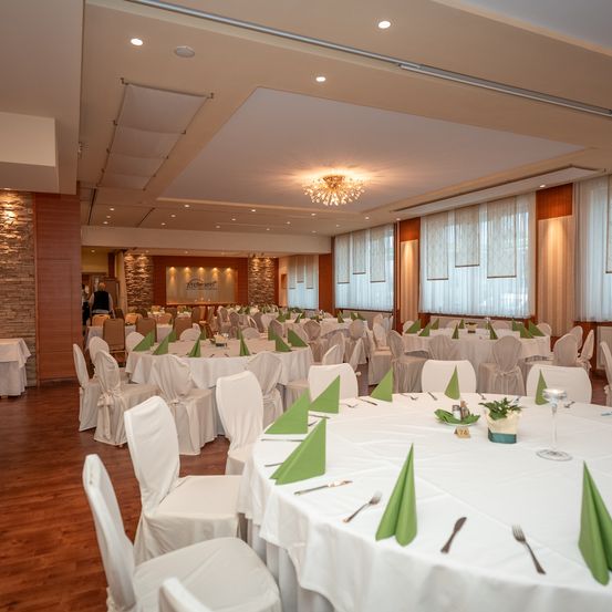 Ein Bankettsaal mit vielen Tischen und Stühlen. Alle Tische sind mit weißen Tüchern und grünen Servietten bedeckt. Der Raum hat einen Holzboden und weiße Wände.