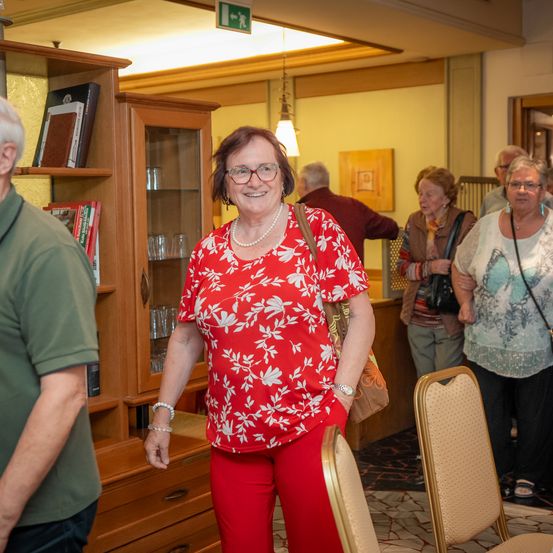Eine Gruppe von Menschen steht in einem Raum mit Holzmöbeln. Eine lächelnde Frau in einem roten Blumenshirt und roten Hosen steht in der Mitte. Andere Menschen stehen hinter ihr, einige tragen Brillen.