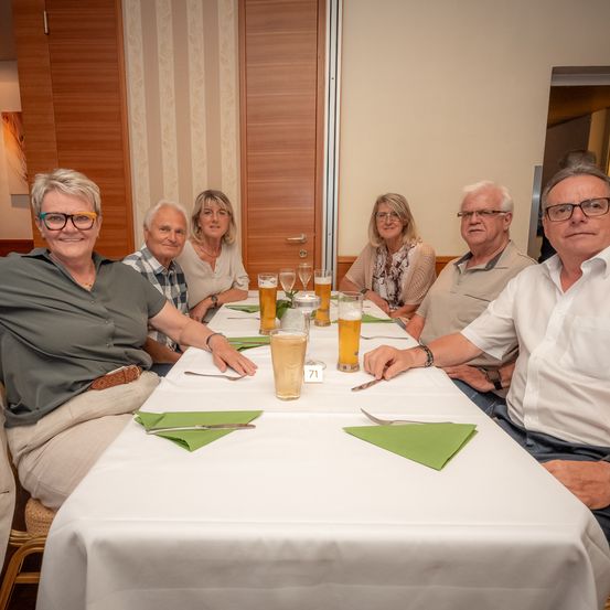 Sechs Erwachsene, alle mit Brille, sitzen um einen Tisch. Sie lächeln und posieren für ein Foto. Der Tisch ist mit einer weißen Tischdecke bedeckt und hat Gläser mit Bier, Besteck und Servietten. Die Nummer 71 steht auf dem Tisch. Der Hintergrund zeigt eine Holztür.