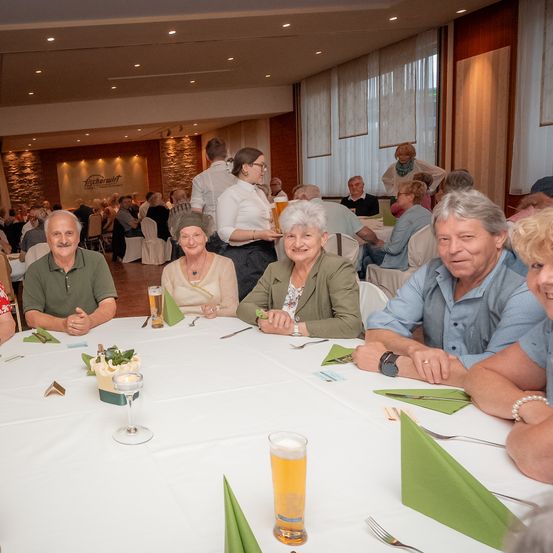 Eine Gruppe von Menschen sitzt an einem Tisch in einem Restaurant, lächelt und trinkt Bier. Eine Frau in einer grünen Jacke steht in der Nähe.