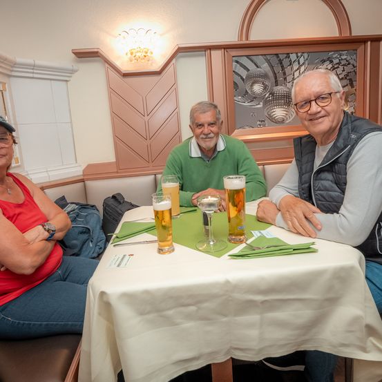 Drei Personen sitzen an einem Tisch in einem Restaurant mit Biergläsern und einer Tasche. Einer trägt eine Uhr und einen Hut, ein anderer ein grünes Shirt und der dritte eine graue Weste.