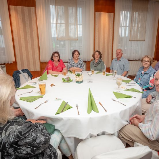 Eine Gruppe älterer Menschen sitzt lächelnd um einen runden Tisch in einem Esszimmer. Der Tisch ist mit grünen Servietten, Besteck und Getränken gedeckt. Hinter ihnen befinden sich weiße Vorhänge und Holzwände.