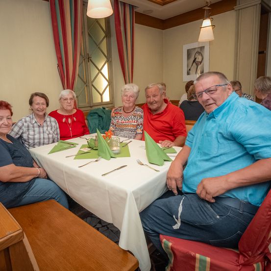 Eine Gruppe älterer Menschen sitzt um einen Tisch in einem Restaurant. Der Tisch ist mit einem weißen Tuch bedeckt. Sie lächeln und posieren für ein Foto.
