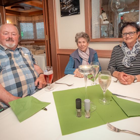 Drei Menschen sitzen lächelnd an einem Tisch in einem Restaurant. Der Tisch hat Weingläser, Teller und grüne Servietten. Hinter ihnen steht ein Schwarzes Brett mit Weinpreisen.