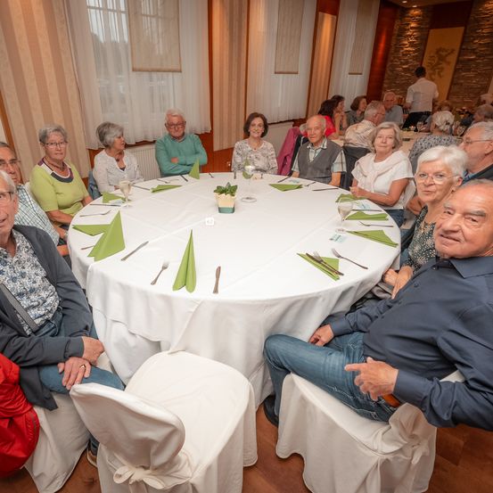 Eine Gruppe älterer Menschen sitzt um einen runden Tisch mit einer weißen Tischdecke, grünen Servietten und Besteck herum. Sie scheinen eine Versammlung oder ein Treffen zu haben.