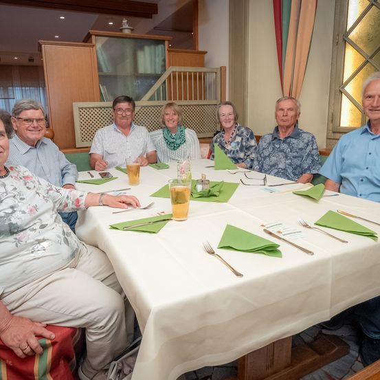 Eine Gruppe von Menschen, alle lächelnd, sitzt um einen Tisch in einem Restaurant. Jede Person hat ein Glas Bier und eine grüne Serviette. Der Tisch ist mit Besteck gedeckt und sie sind von einer Holzwand umgeben.