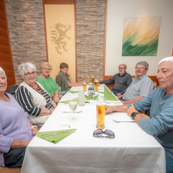 Eine Gruppe älterer Menschen sitzt um einen langen Tisch in einem Restaurant. Gläser und Getränke stehen auf dem Tisch, und der Raum hat Steinwände und ein Gemälde.