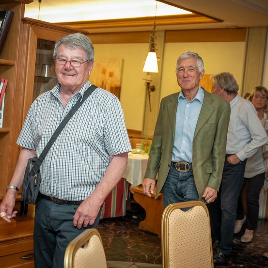 Zwei ältere Männer stehen in einem Raum mit einem Holzschrank, einem Tisch und Stühlen. Einer trägt ein kariertes Hemd und eine Umhängetasche, der andere einen grünen Blazer. Hinter ihnen stehen zwei weitere Personen, eine in einem weißen Hemd und die andere in einem Kleid.