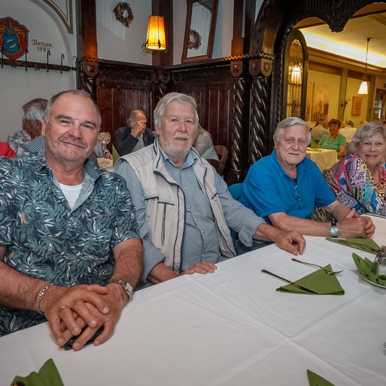 Eine Gruppe älterer Männer und Frauen sitzt um einen Tisch herum, wahrscheinlich in einem Restaurant. Sie lächeln und scheinen ihre Mahlzeit zu genießen.