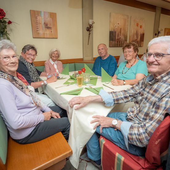 Eine Gruppe älterer Menschen sitzt an einem Tisch, lächelt und schaut in die Kamera. Sie haben eine Tischdecke und Blumen auf dem Tisch.