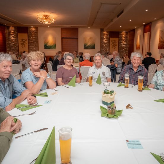 Eine Gruppe älterer Menschen sitzt um einen Tisch mit Getränken und einem Kuchen herum in einem Essbereich mit weißen Tischdecken, Stühlen und dekorativen Elementen an den Wänden.