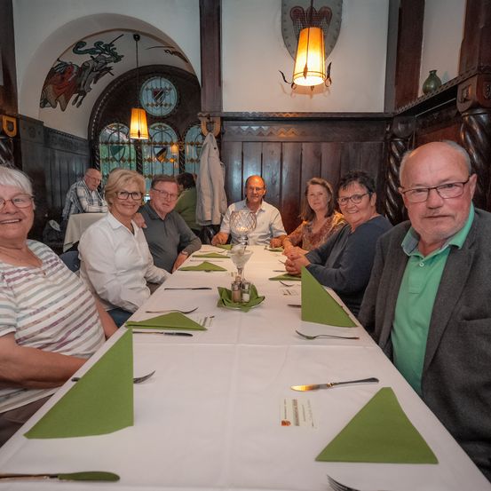 Eine Gruppe von Menschen sitzt an einem langen Tisch in einem traditionellen deutschen Restaurant. Der Tisch ist mit grünen Servietten, Gabeln und Messern gedeckt. Das Restaurant hat Holzwände und gewölbte Fenster mit Buntglas.