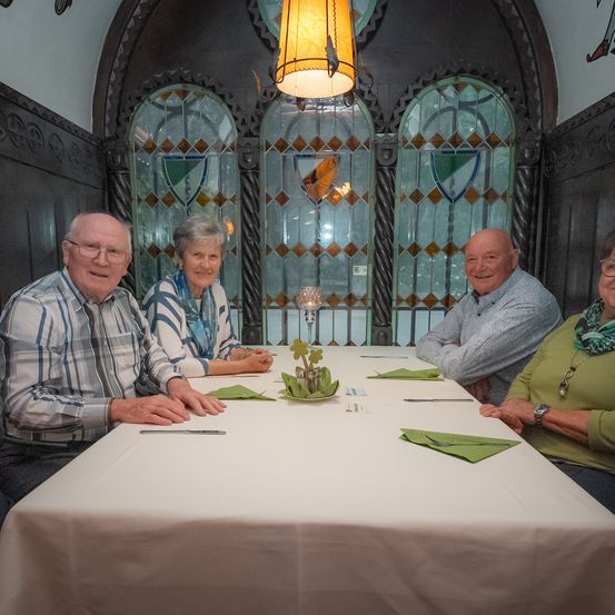 Vier Personen sitzen um einen Tisch in einem gemütlichen, schwach beleuchteten Raum. Der Tisch ist mit einer weißen Tischdecke und grünen Servietten bedeckt. Der Raum hat bogenförmige Buntglasfenster.