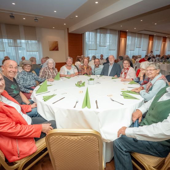 Eine Gruppe älterer Menschen versammelt sich an einem runden Tisch in einem Speisesaal. Der Tisch ist mit einer weißen Tischdecke und grünen Servietten bedeckt. Sie lächeln und schauen zur Kamera. Der Raum hat eine weiße Decke und Wände.