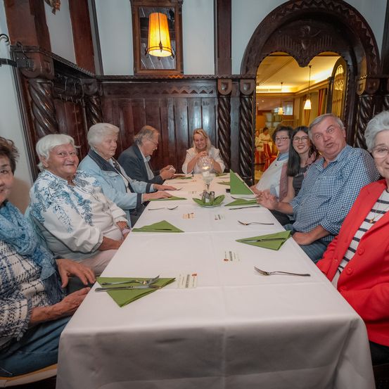 Eine Gruppe älterer Menschen sitzt in einem Restaurant um einen langen Tisch herum, lächelt und plaudert. Sie tragen Freizeitkleidung und haben verschiedene Gegenstände auf dem Tisch, darunter Besteck, Servietten und Gläser.