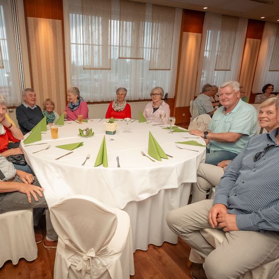 Eine Gruppe älterer Menschen sitzt um einen runden Tisch in einem Esszimmer. Der Tisch ist mit weißen Tischdecken, Weingläsern und grünen Servietten gedeckt. Der Raum hat Holzböden und große Fenster mit weißen Vorhängen.