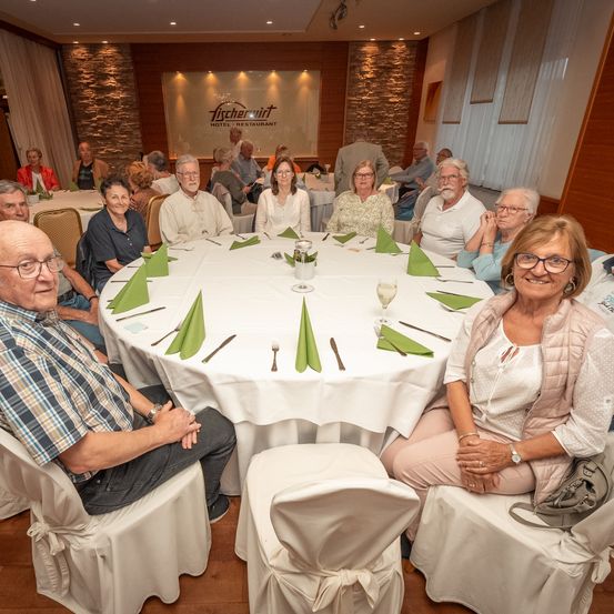 Eine Gruppe von Menschen sitzt um einen runden Tisch in einem Restaurant. Der Tisch ist mit grünen Servietten und Besteck gedeckt. Ein Schild im Hintergrund lautet Fischerwirt Hotel Restaurant.