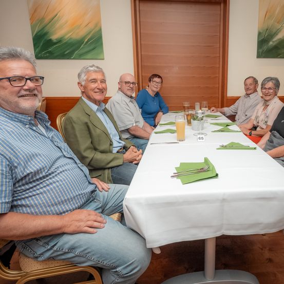 Sieben Menschen sitzen lächelnd um einen langen Tisch in einem Esszimmer. Ein Tisch hat Gläser, eine Nummer 65 und grüne Servietten. An der Wand hängen Gemälde.