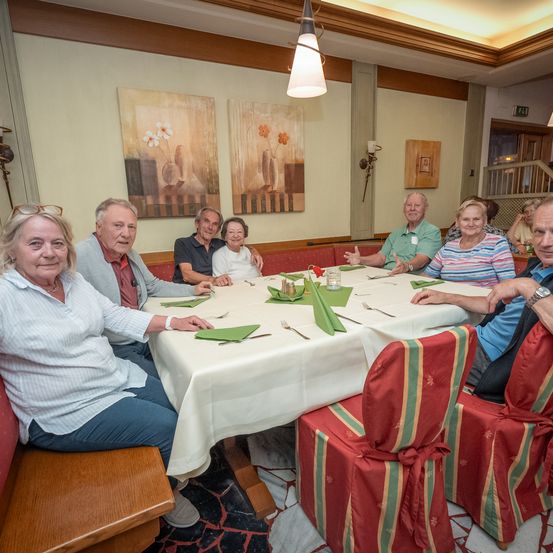 Eine Gruppe älterer Menschen sitzt um einen Tisch herum, wahrscheinlich in einem Restaurant. Sie lächeln und posieren möglicherweise für ein Foto. Der Tisch ist mit weißer Tischdecke, Besteck und grünen Servietten gedeckt. Der Raum hat weiße Wände und Gemälde darauf.