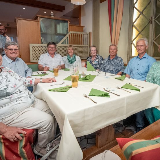 Eine Gruppe von Menschen sitzt um einen Tisch herum und lächelt für ein Foto. Der Tisch ist mit grünen Servietten und Besteck gedeckt. Ein Mann mit Brille hält ein Glas Bier. Eine Frau in einem Blumenhemd sitzt links, und ein Mann mit Bart steht hinter dem Tisch.