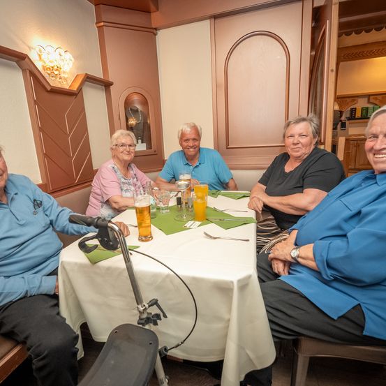 Fünf ältere Erwachsene sitzen in einem Restaurant um einen Tisch. Sie haben Bier und lächeln. Der Tisch hat eine weiße Tischdecke und grüne Tischsets.