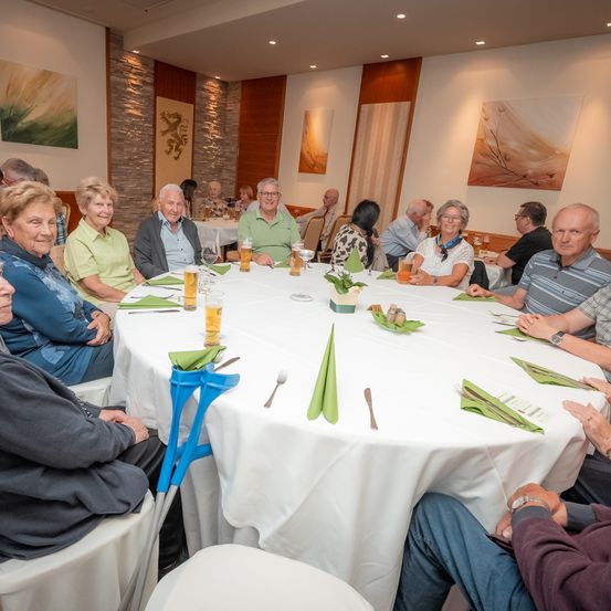 Eine Gruppe älterer Erwachsener sitzt um einen Tisch in einem Esszimmer, einige mit Getränken und Essen. Der Tisch ist mit einer weißen Tischdecke bedeckt.