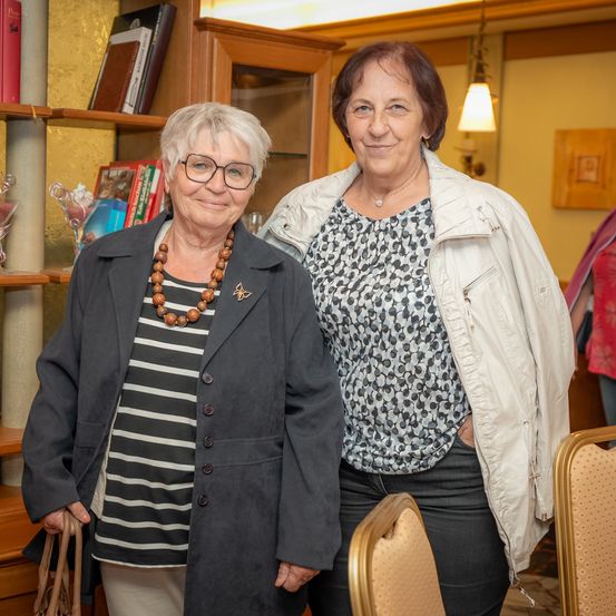 Zwei Frauen stehen nebeneinander in einem Raum mit Holzmöbeln, lächeln und posieren für ein Foto.