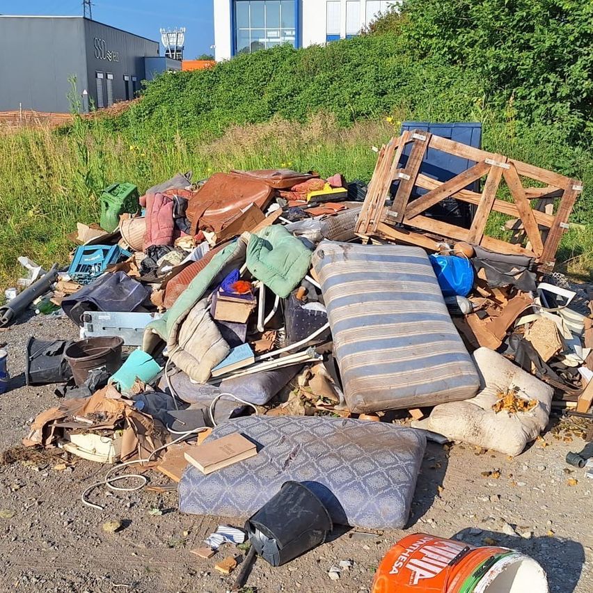 Ein vermüllter Bereich mit verschiedenen Gegenständen, einschließlich Matratzen, Eimern, Holzpaletten und anderem Schutt, verstreut auf dem Boden in der Nähe eines Gebäudes.