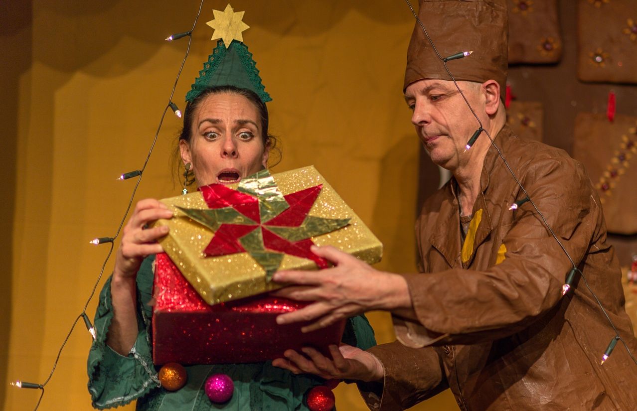 Eine Frau in einer Weihnachtskostüm hält ein verpacktes Geschenk, während ein Mann in einem braunen Hut zusieht. Sie tragen beide Weihnachtslichter auf ihren Hüten.