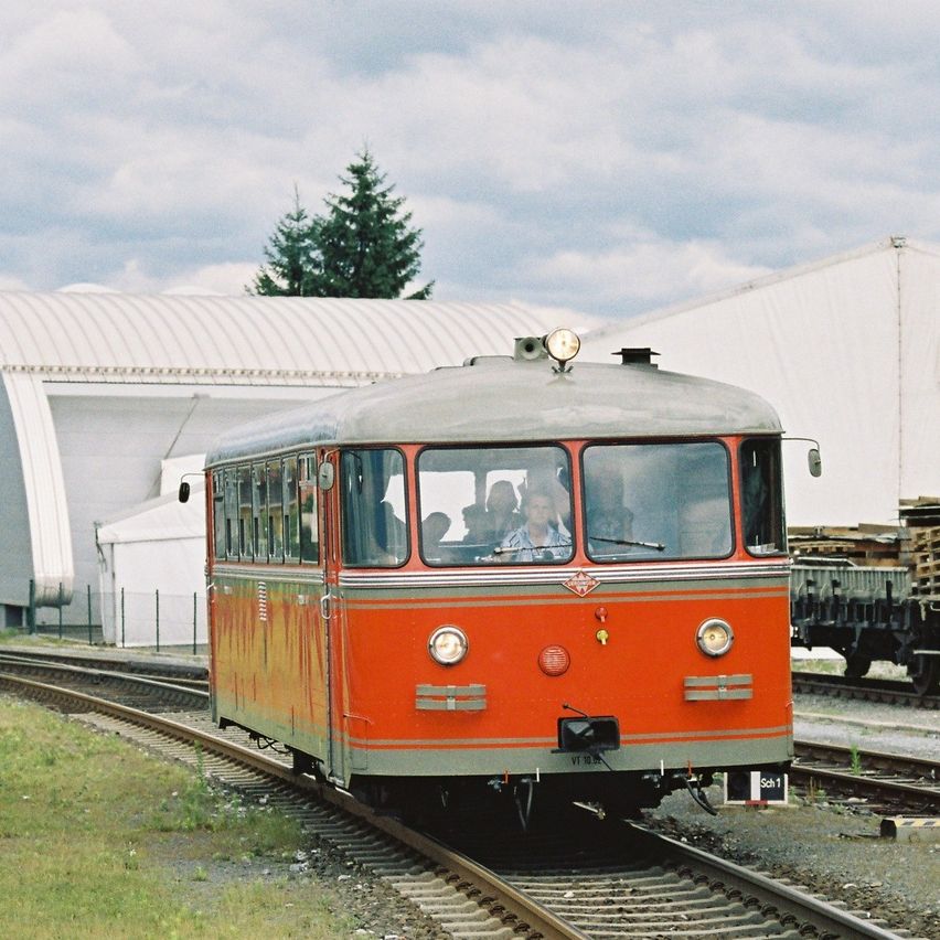 Ein altmodischer roter Zug fährt auf einer Eisenbahnstrecke, mit Passagieren im Inneren und einem weißen Gebäude im Hintergrund.
