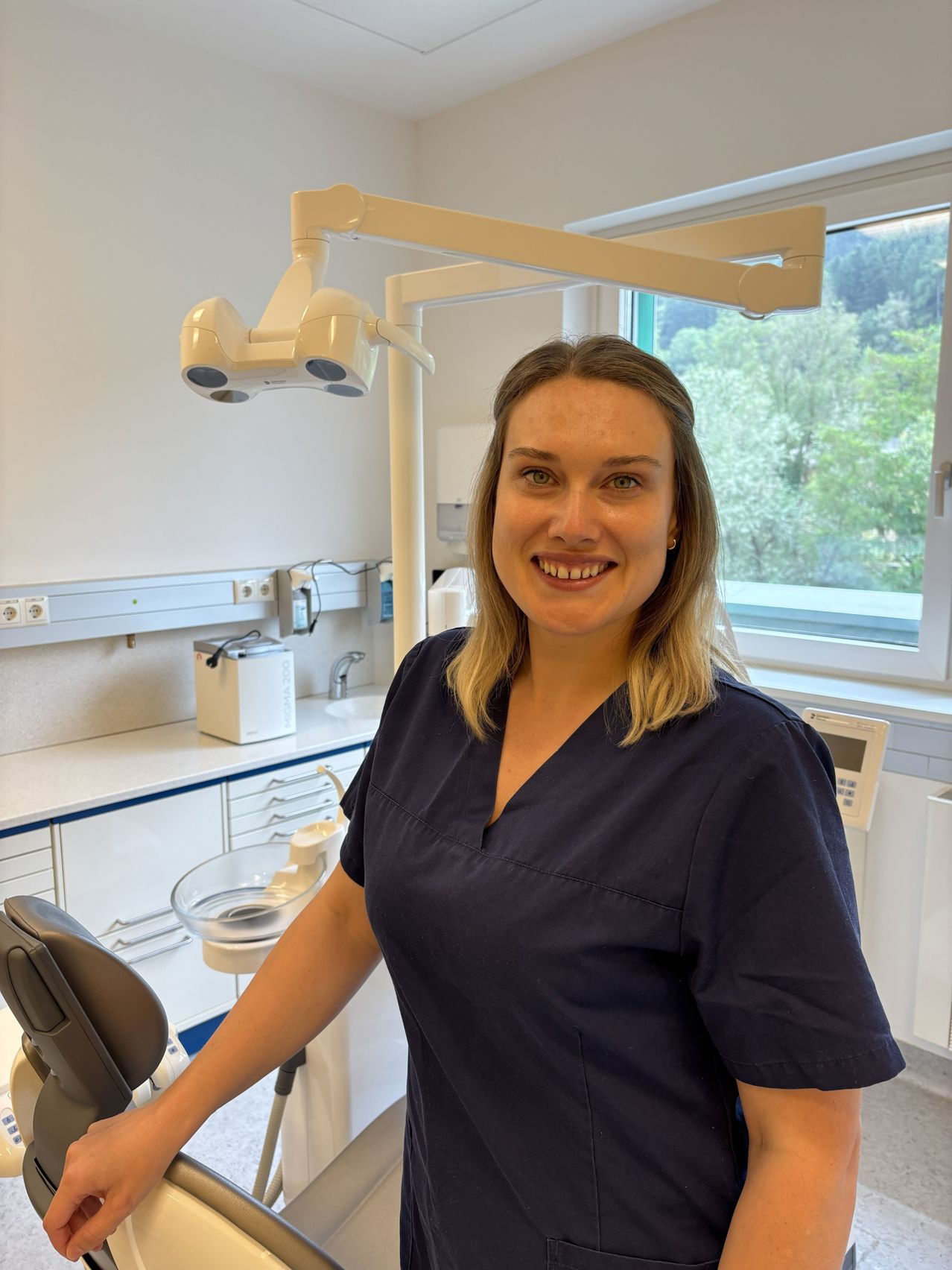 A woman in a blue scrub suit is standing in a dental clinic with a smile, near a dental chair and a window with trees outside.