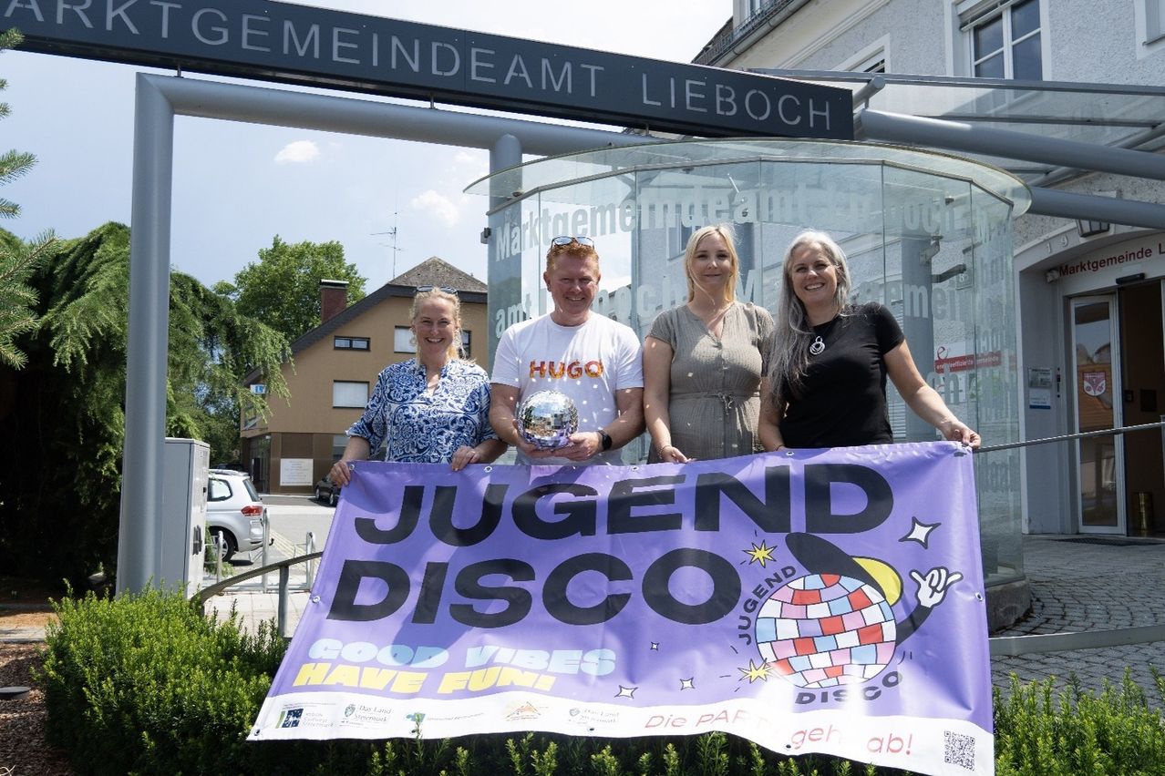 Four people pose with a banner and disco ball in front of a sign for Jugend Disco. The sign reads Jugend Disco with Good Vibes Have Fun.