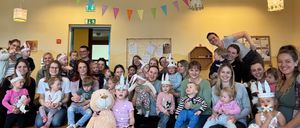 Eine Gruppe von Menschen lächelt und posiert für ein Foto in einem Raum. Einige Leute sitzen auf dem Boden und ein Plüschtier steht in der Mitte. Einige bunte Flaggen hängen an der Decke. Ein Brett ist an der Wand montiert und einige Lampen sind an der Decke montiert.